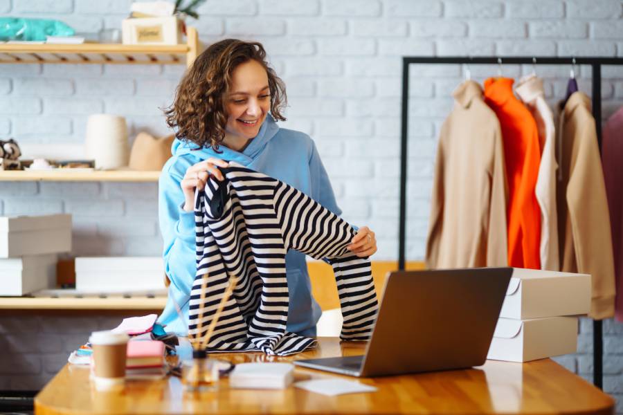 Vendedora de moda revisando una prenda frente a su laptop en una tienda, representando cómo una buena gestión mejora la experiencia de compra del cliente