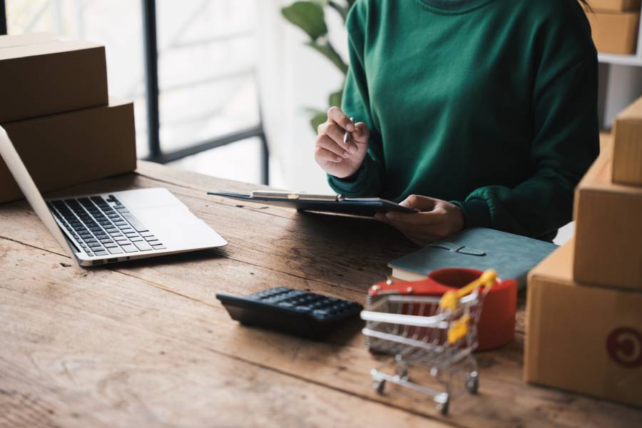 Persona con suéter verde anotando en una tabla con portapapeles, rodeada de cajas de envío, una laptop, una calculadora y un carrito de compras en miniatura, gestionando operaciones en una plataforma de marketplace