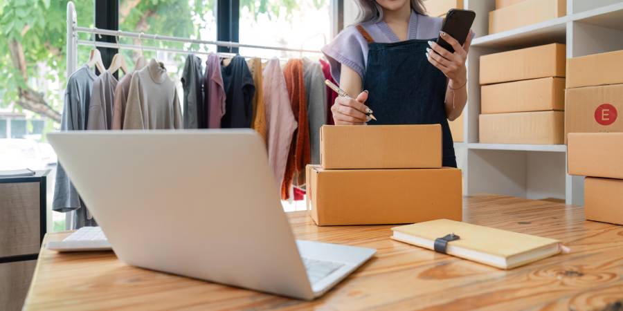 
Vendedora revisando pedidos con celular y laptop en tienda de ropa, gestionando ventas en marketplace desde su negocio