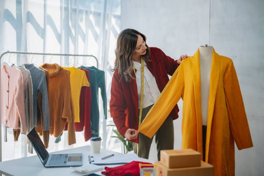 Diseñadora midiendo prenda amarilla en maniquí rodeada de fotos de productos de moda para vender en Mercado Libre. 