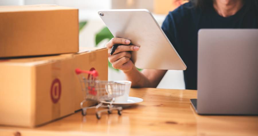 Trabajador de ecommerce revisando tablet junto a cajas de envío, laptop y carrito de compras en escritorio 