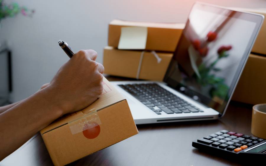 Persona escribiendo en caja de envío junto a laptop y calculadora, gestionando pedidos en plataforma de e-commerce 